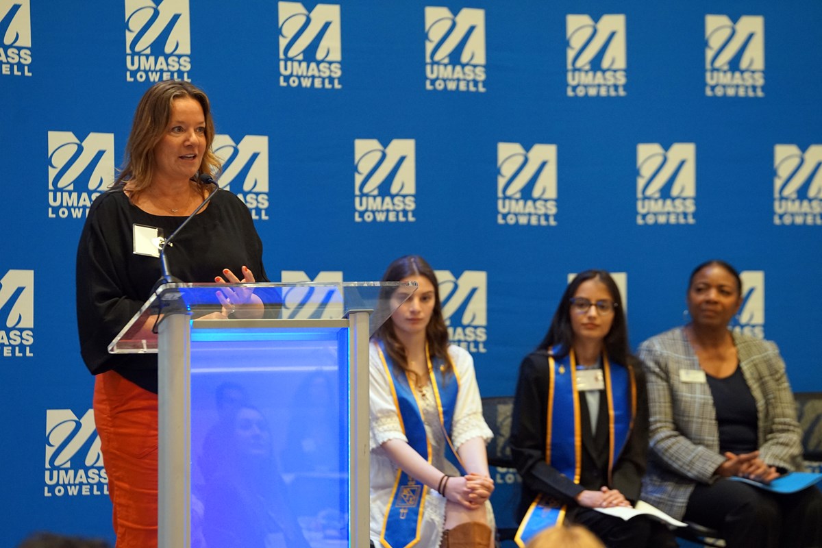 A person speaks at a podium while three people are seated behind her on a stage.